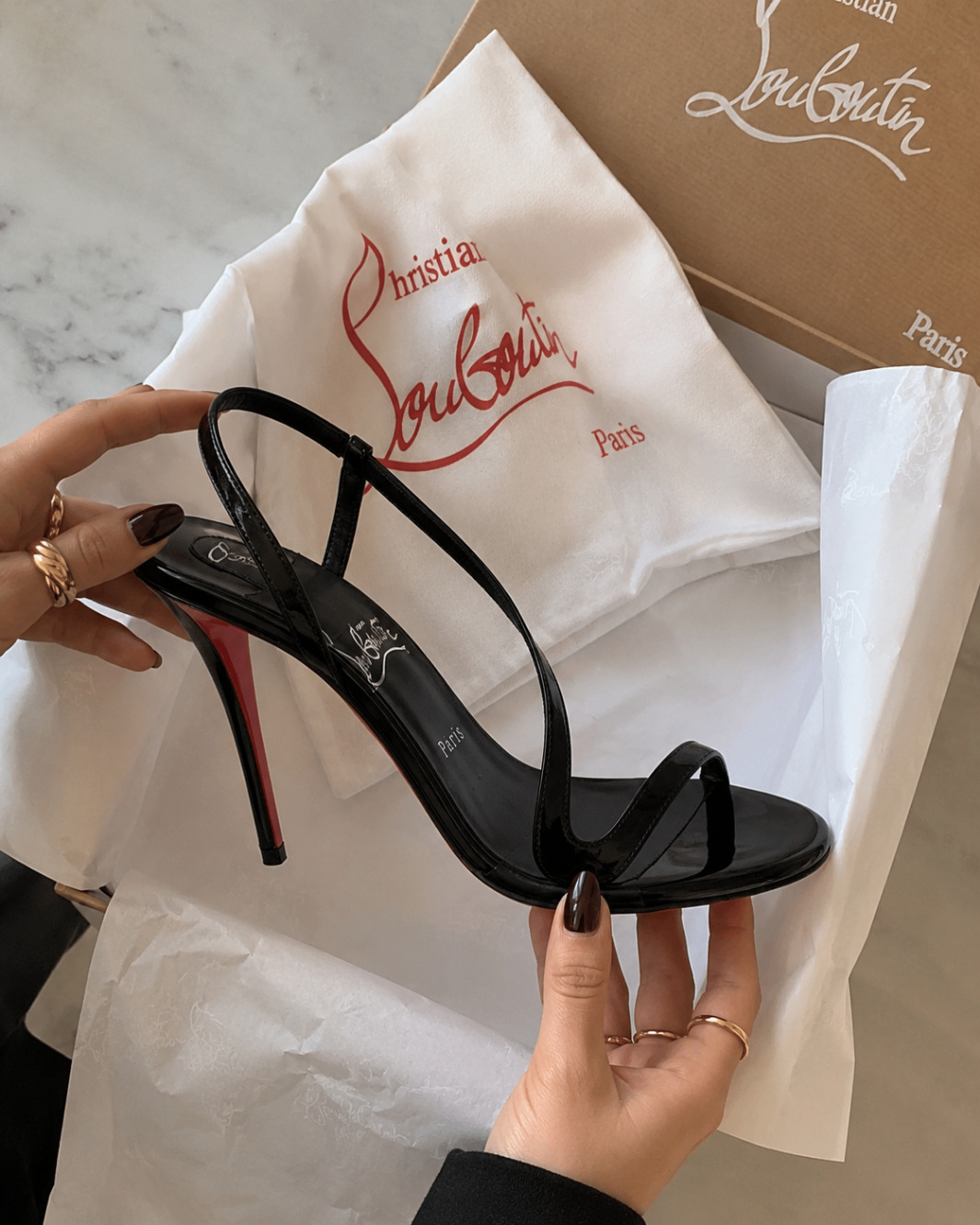 Hands holding a black patent strappy stiletto sandal above its tissue-lined box